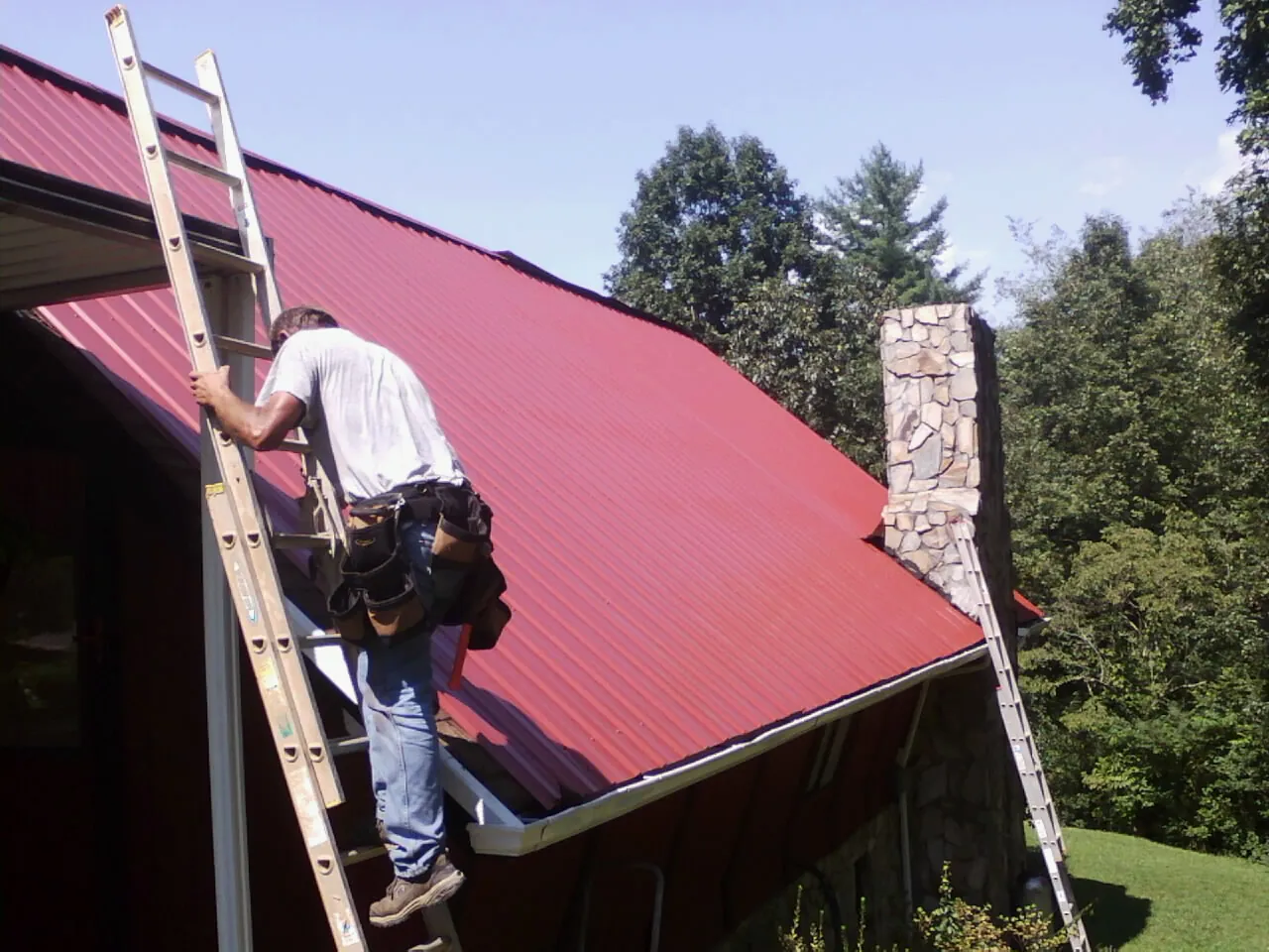 Roofer on a ladder beside a red metal roof and stone chimney — Soffit & Fascia Repair by Roofing Brothers in Xenia